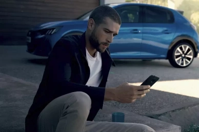 Homme utilisant l'application My Peugeot sur son smartphone devant une Peugeot 208 bleue.